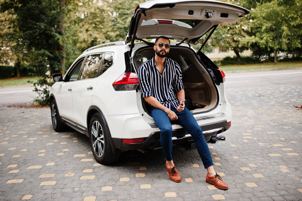 Successful arab man wear in striped shirt and sunglasses sitting in the trunk of his white suv car with mobile phone. - Fotó, kép