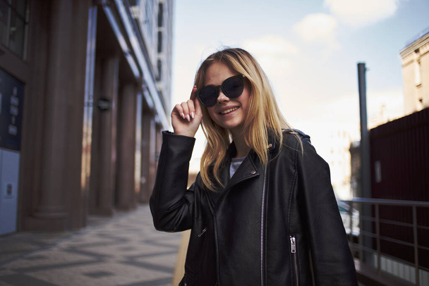 Donna alla moda in giacca di pelle e occhiali da sole vicino all'edificio sulla strada - Foto, immagini