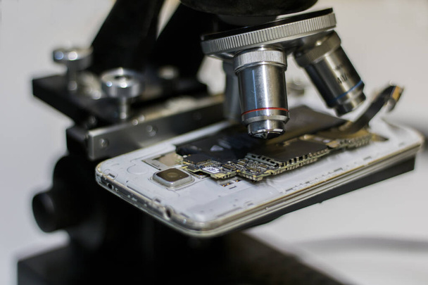 close-up zijaanzicht van een microscoop, terwijl het analyseren van de circuits op de achterkant van een smartphone. - Foto, afbeelding