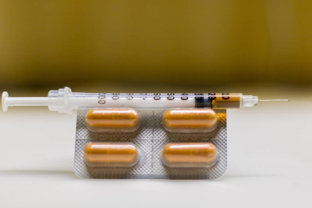 Frontal view in the foreground, of a package of drugs in orange capsules, with a syringe full of liquid cocaine positioned on top, all placed on a white and shiny surface. - Photo, Image