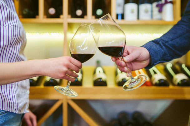 Close up front view on male and female caucasian hands holding glasses of red wine toasting in front of the shelf man and woman celebrating concept - Photo, Image