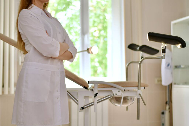 A doctor in white uniform with folded hands. Stands in profile in front of a gynecological chair with footrests. In the medical office of the clinic. - Foto, Imagem