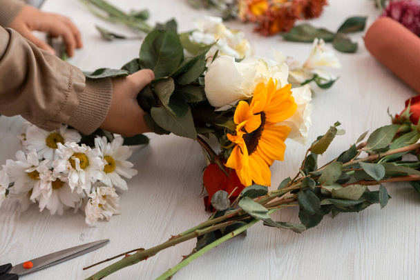 Floristin bastelt mit ihren Händen ein Blumengesteck mit Sonnenblumen, Rosen und anderen Blumen auf einem weißen Tisch, Beruf - Foto, Bild