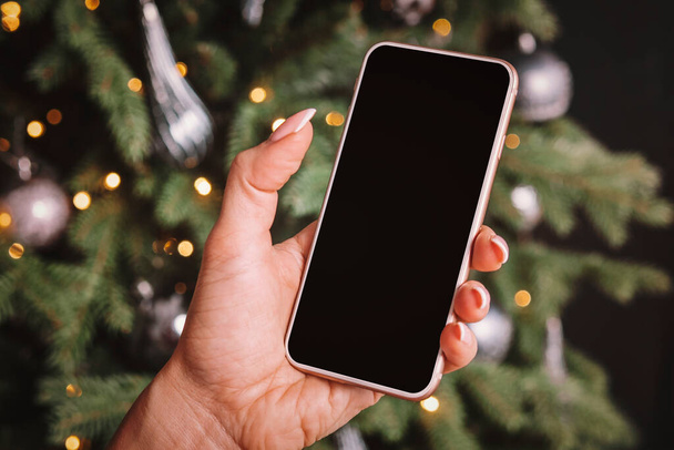 Smartphone στο χέρι μιας γυναίκας στο φόντο ενός χριστουγεννιάτικου δέντρου. Κενό διάστημα στην οθόνη για σχεδιασμό και κείμενο. - Φωτογραφία, εικόνα
