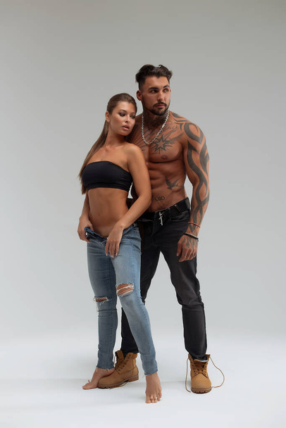 Attractive female embracing handsome shirtless male with tattoos from behind and looking at camera against gray background - Photo, Image