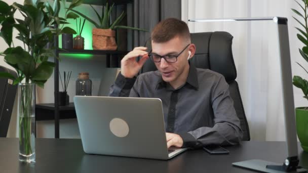 Smiling caucasian man wearing earphones is doing online video chat conference sitting at office desk and looking at laptop computer. Businessman is making meeting conference webinar from office - Záběry, video