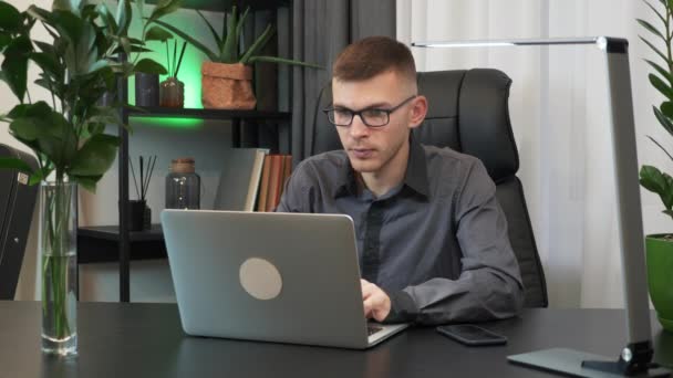 Motivated young man is closing laptop computer and finishing to work at workplace desk in the evening. Businessman in glasses finishes his work on notebook in office. Business concept - Imágenes, Vídeo
