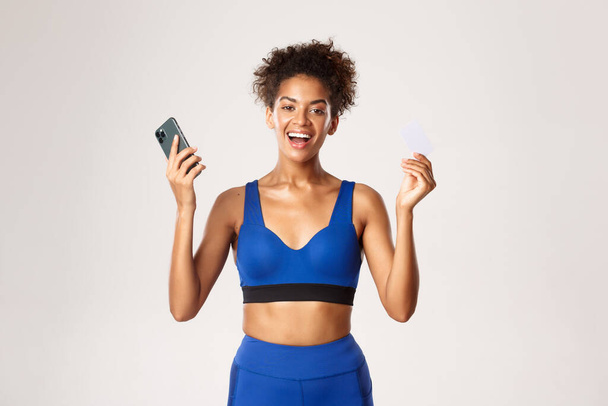 Neşeli Afro-Amerikan bayan sporcu, neşeli ve gülümseyen, akıllı telefon ve kredi kartı tutan, beyaz arka planda duran - Fotoğraf, Görsel