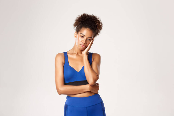 Photo studio d'une athlète afro-américaine désabusée et ennuyée en tenue de sport bleue, penchée sur la main et regardant la caméra s'ennuyer, debout sur fond blanc - Photo, image