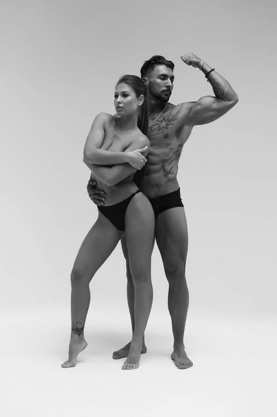 Black and white side view of topless woman and shirtless man looking at camera against gray background - Foto, immagini