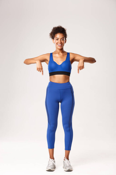 Volledige lengte van mooie Afrikaans-Amerikaanse fitness meisje, het dragen van sportkleding, wijzende vingers naar beneden en glimlachen met gelukkig gezicht, tonen van reclame of logo, witte achtergrond - Foto, afbeelding