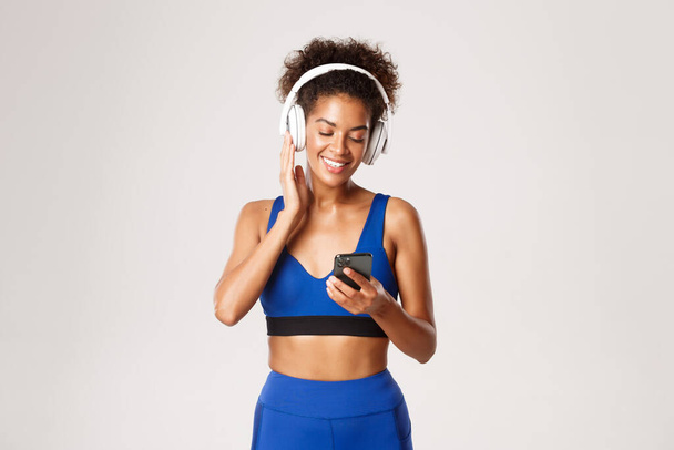 Beautiful fitness woman in blue sportswear, workout with music in wireless headphones, using smartphone application, standing against white background - Foto, Imagen