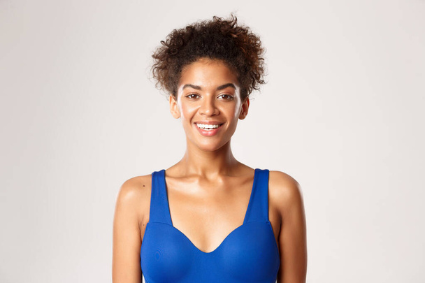 Koncepcja treningu i sportu. Zbliżenie zdrowej i wysportowanej afrykańsko-amerykańskiej kobiety fitness w niebieskiej odzieży sportowej, uśmiechnięta szczęśliwa przed kamerą, stojąca na białym tle - Zdjęcie, obraz