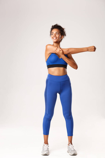 Full length of smiling beautiful african-american sportswoman, stretching hands, warming-up before workout and looking left determined, standing over white background - Photo, Image