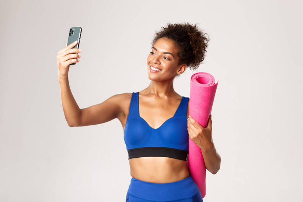 Young beautiful african-american fitness woman in sport outfit, taking selfie with yoga mat, taking photo on smartphone, standing against white background - Photo, image