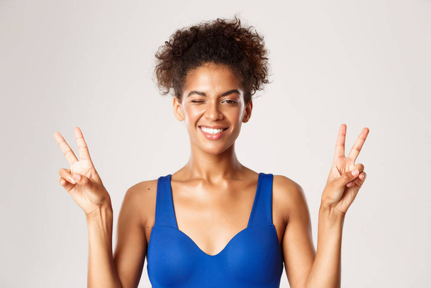 Cheerful african-american fitness girl in blue workout clothing, showing peace signs and smiling happy, standing over white background - Photo, Image