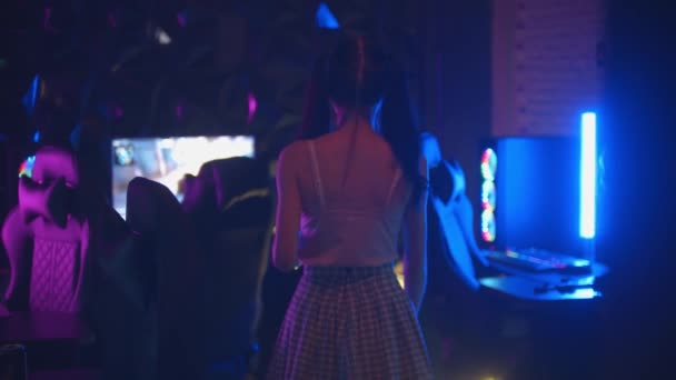 Giovane donna sexy con code di cavallo cammina nel club di gioco in piedi accanto alla sedia e tenendo un joystick - Filmati, video