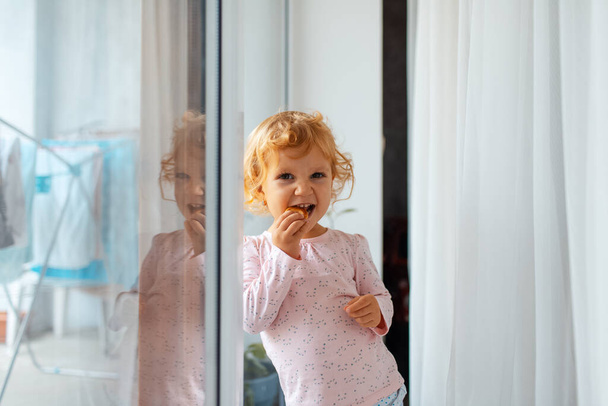 窓の近くの家でベーグルを食べる子供の自然な肖像画. - 写真・画像
