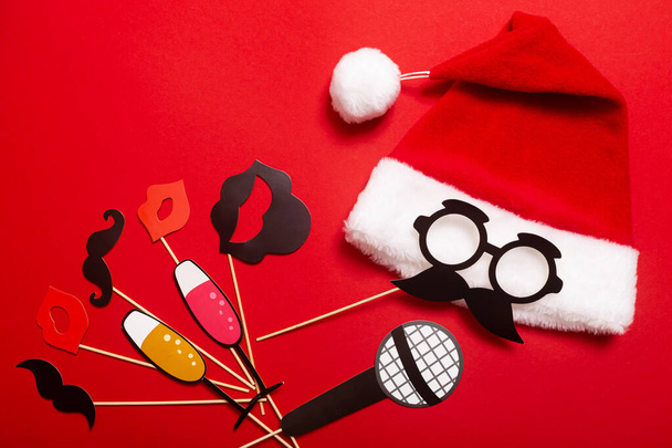 Rotes Banner mit Weihnachtsmütze mit weißem Bommel und einem Gesicht aus Brille und Schnurrbart. Requisiten für die Unterhaltung der Gäste bei einem Neujahrsfest, Speed-Dating oder einer Karaoke-Party. - Foto, Bild
