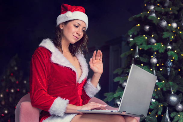 mujer joven con un disfraz de Papá Noel se sienta frente a una pantalla de computadora y agita sus manos. Saludos de Navidad en línea. Años nuevos en cuarentena. - Foto, imagen