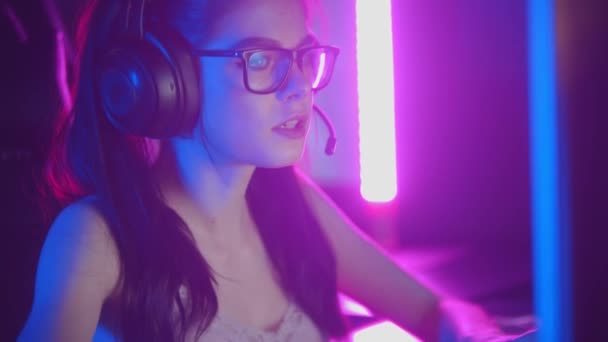 Νεαρή γυναίκα με γυαλιά που παίζει online παιχνίδια στο νέον gaming club - φορώντας ακουστικά και μιλώντας στο μικρόφωνο - Πλάνα, βίντεο