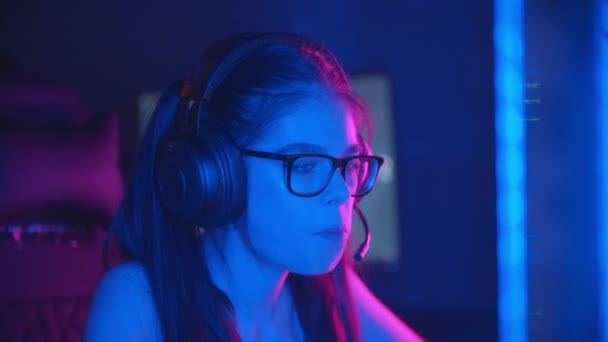 Gözlüklü genç bir kadın modern neon oyun kulübünde online oyunlar oynuyor. Kulaklık takıyor ve mikrofonda konuşuyor. - Video, Çekim