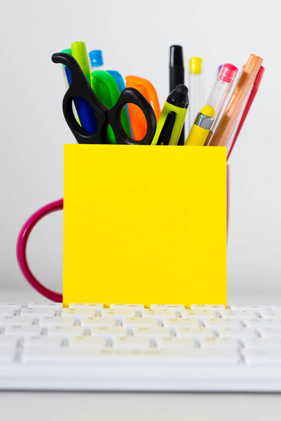 Tastiera per computer, cartellino giallo vuoto con spazio per il testo, appoggiato a una tazza piena di pennarelli, pennarelli, matite e forbici. - Foto, immagini