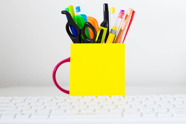Teclado do computador, um cartão amarelo em branco com espaço para texto, encostado a uma xícara cheia de canetas, canetas de feltro, lápis e tesoura. - Foto, Imagem