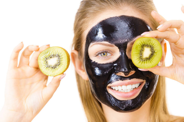 Young woman with carbo black peel-off mask on her face holding kiwi fruit halves, covering eyes, on white. Beauty treatment. Skincare. - Foto, Imagen