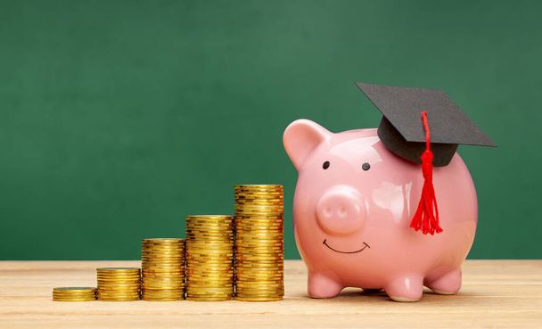 Čuňasova banka v maturitním klobouku poblíž hromady mincí. Úspory na vzdělání. Ceny vysokoškolského vzdělávání. Zelená školní rada pozadí - Fotografie, Obrázek