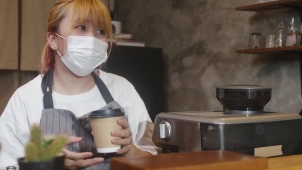Young Asia female barista wear face mask serving take away hot coffee paper cup to consumer at cafe. Owner small business, lifestyle new normal after corona virus and social distancing concept. - Footage, Video