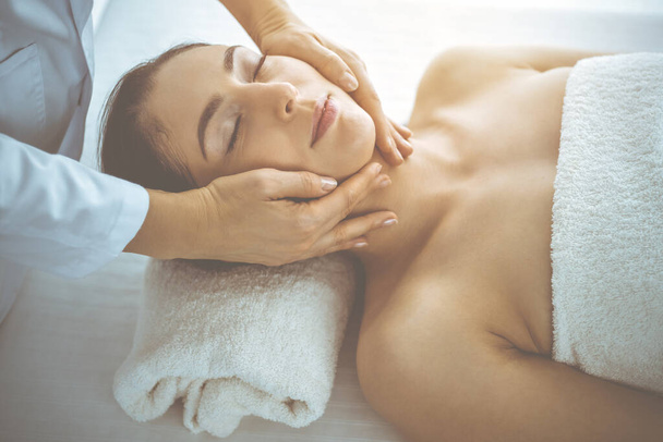 Beautiful brunette woman enjoying facial massage with closed eyes in sunny spa center. Relaxing treatment and cosmetic medicine concepts - Foto, Bild
