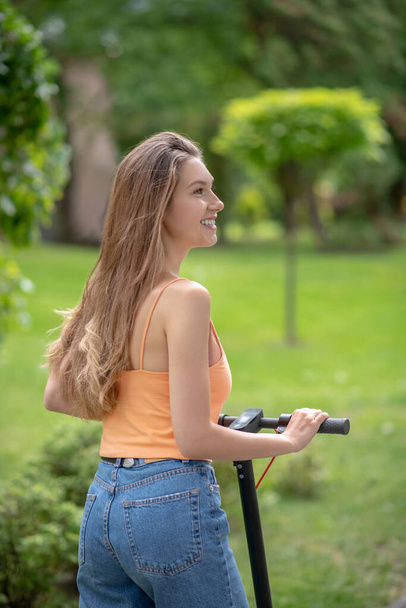 Long-haired young girl in jeans riding a scooter in the park - Zdjęcie, obraz