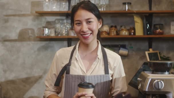 Muotokuva nuori aasialainen nainen barista tarjoilija tilalla kahvikuppi onnellinen kaupunkien kahvilassa. Aasia pienyrittäjä tyttö esiliina rentoutua hampainen hymy haluavat kamera seistä laskuri kahvilassa. - Materiaali, video