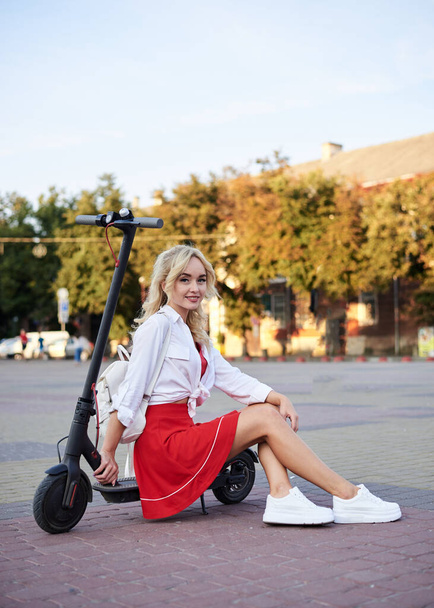 Young blond woman, sitting on black electric scooter in city center in front of old historical building with pillars. Summer leisure activity. Spending free time outdoors when weather is warm. - Фото, изображение