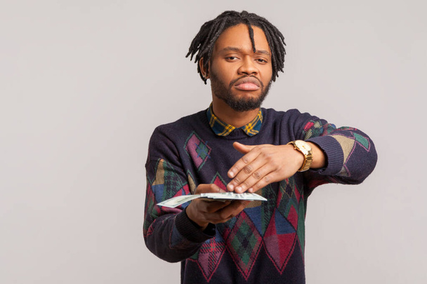 Impudent rich african man with dreadlocks in casual style sweatshirt scattering dollars with self confident face, wasting money. Indoor studio shot isolated on gray background - 写真・画像