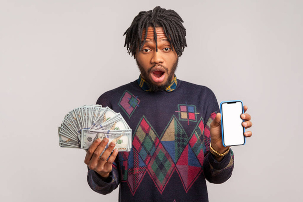 Shocked african man with dreadlocks and beard looking at camera with big eyes and open mouth, holding empty blank smartphone and dollars in hands - Fotografie, Obrázek