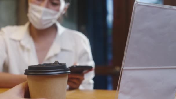 A fiatal ázsiai nők arcmaszkot viselnek, önkiszolgáló mobiltelefonos fizetést használnak érintkezés nélkül az étteremben. Lány barista beszél kap megrendelést az ügyfél a kávézóban. Életmód új normális után koronavírus koncepció. - Felvétel, videó