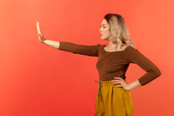 Side view portrait of serious young woman with hand on the waist in casual clothes demonstrate stop gesture by her hand. Forbidden and decline concept. Indoor studio shot isolated on red background - Foto, Imagen