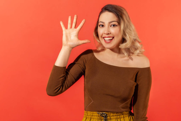Hello! Portrait of beautiful smiling young lady with blonde hair in brown blouse with raised hand demonstrate hello gesture and ready to make friendship. Indoor studio shoot isolated on red background - Foto, immagini