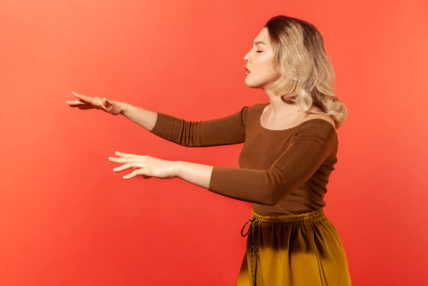 Side view portrait of blonde beautiful woman in brown blouse standing with closed eyes and outstretched hands, trying to move forward blind. Indoor studio shot isolated on red background - Zdjęcie, obraz
