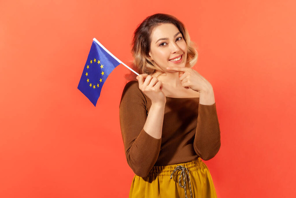 Happy smiling woman pointing finger at europe union blue flag with yellow stars, open borders for traveling. Indoor studio shot isolated on orange background - Foto, imagen