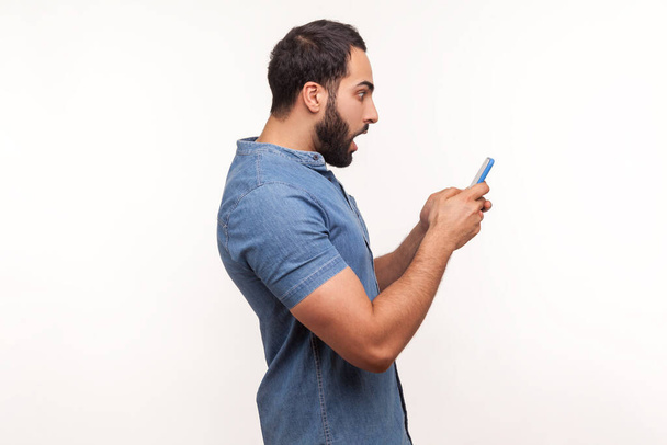 Profile portrait bearded man in shirt looking at smartphone display with widely open mouth, shocked and surprised with application. Indoor studio shot isolated on white background - Photo, image