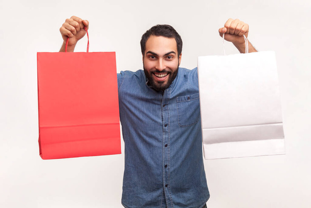 Positive happy man in blue shirt showing two paper bag looking at camera with toothy smile, successful shopping. Indoor studio shot isolated on white background - Photo, Image