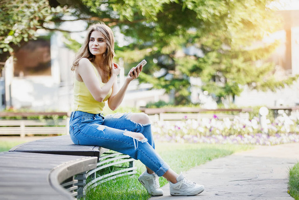 Vrouw met smartphone in geel T-shirt en jeans in een stadspark. - Foto, afbeelding