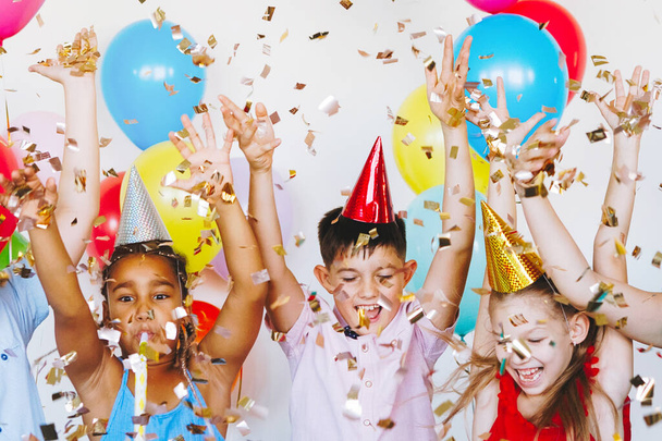 Яркие, милые дети празднуют день рождения. Многонациональная вечеринка, воздушные шары, конфетти, кепки, улыбки, подростки счастливы поднимать руки. - Фото, изображение