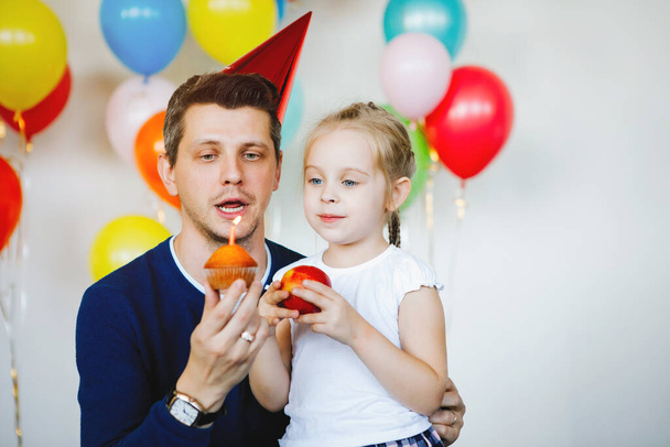 Baba ve kızı doğum günü pastasını mumla kutluyorlar. Kırmızı şapkalı baba, mavi gözlü sarışın, at kuyruklu ve kırmızı elmalı kız. Aile mutluluğu, mutlu gülümseme, babalık sevinci.. - Fotoğraf, Görsel