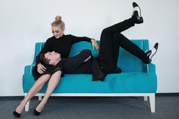 Cool mec en costume noir et bottes blanches se trouve sur les genoux d'une fille blonde sur un canapé turquoise. Homme et femme sur le canapé. - Photo, image