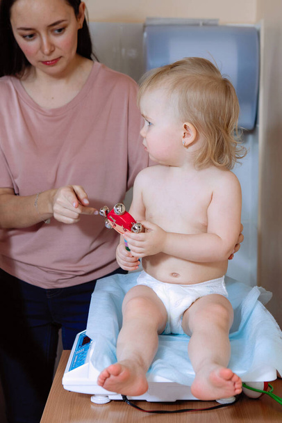 Bébé blonde assise sur la balance dans une clinique médicale. A proximité se trouve une mère attentionnée. - Photo, image