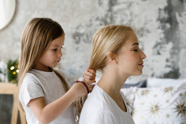 Concept of tender moments. Profile portrait of focused girl braids hair and making hairstyle for her dreamy mother. Charming daughter, spending free time together with mom and playing at home - Fotó, kép